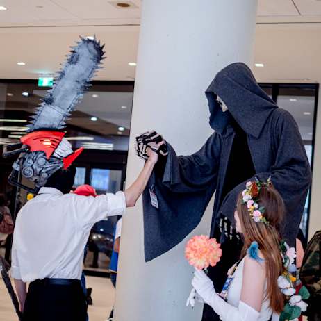 Denji from Chainsaw Man and the Grim Reaper join hands as an onlooker dressed as an anime character watches. 