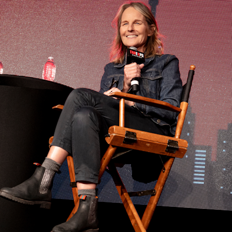 Helen Hunt sits in the director's chair on the main stage holding a FAN EXPO Canada branded microphone. She smiles at the audience and sits cross-legged. She is wearing an all black denim outfit.