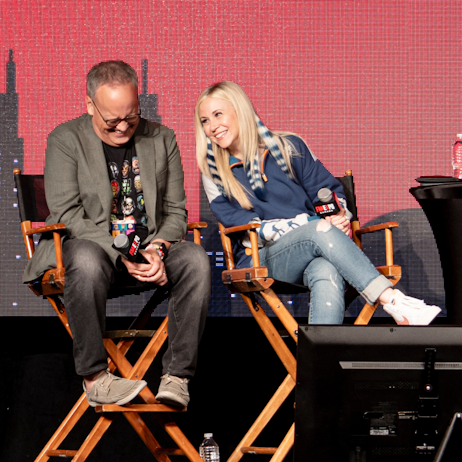 Dee Bradley Baker looks down in laughter, Ashley Eckstein smiles and leans into him. Both are holding FAN EXPO Canada branded microphones and sitting in directors chairs