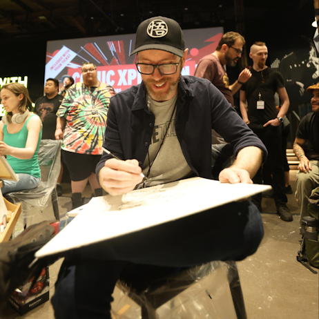 A comic artist leans into his canvas as he outlines his piece. He smiles as he works, and sits with one leg under the other in a chair. Fans watch as he works. 