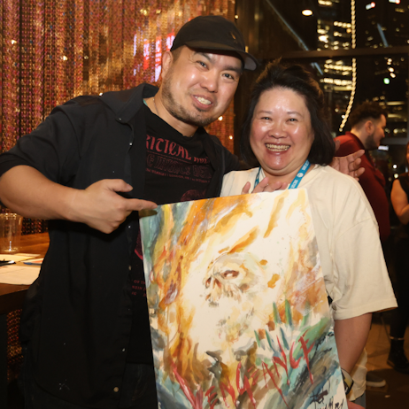 Tony Moy posts with the fan who won the auction for his Ghost Rider painting at Comic Xplosion. Both smile at the camera, and he points proudly at his piece.