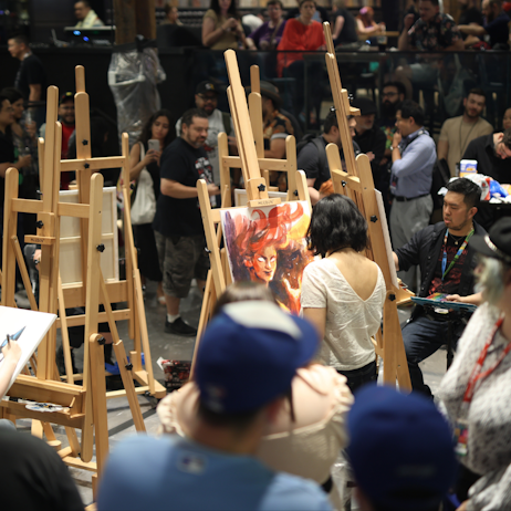 Fans gather around a group of six comic artists painting in the centre of the room. There are six large wooden easels holding up the art as the artists work. 