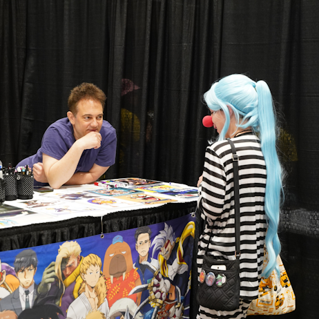 Mike McFarland, in a blue shirt, leans in to listen attentively to a fan at his autograph table dressed as Buggy from One Piece.