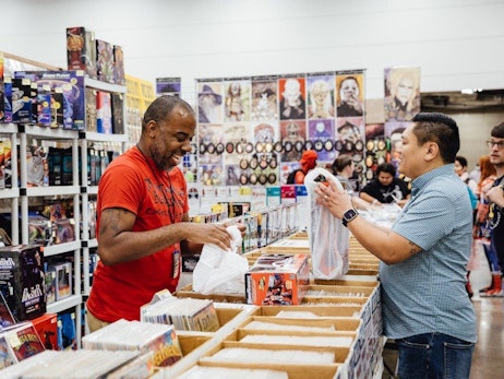Two men browsing at a convention booth filled with comics and merchandise. The man on the left, wearing a red t-shirt with "Comics" written on it, is examining items from a box, while the man on the right, in a blue button-down shirt, looks at a plastic bag containing a purchase. Behind them are shelves stacked with various comic books, toys, and posters. Other attendees can be seen in the background, perusing the offerings.