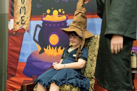 The image captures a young child dressed as a witch, sitting joyfully on an ornate chair at a themed photo booth. The child wears a large, floppy witch's hat and a dark blue dress, with a bright and delighted smile on her face. The backdrop features a colorful illustration of a bubbling cauldron over a fire, enhancing the magical theme. A partial view of another person in a dark green robe, possibly helping with the costume, adds to the playful and festive atmosphere of the setting.