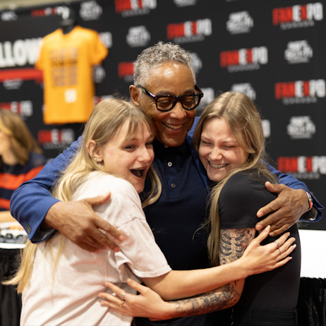 Giancarlo Esposito stands between two fans in a hug. All three smile gleefully at the embrace. They are in front of a table ahead of the black FAN EXPO Canada background. 