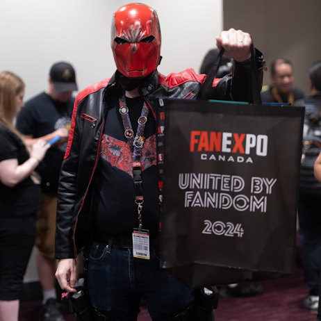 Red hood stands amonst other fans, holding the FAN EXPO Canada 2024 official merchandise tote bag. 