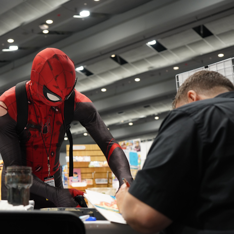 Spider-man approaches the table of a comic artist for an autograph. The artist, whose back is to the camera, signs the comic art (not fully visible) in their hand.