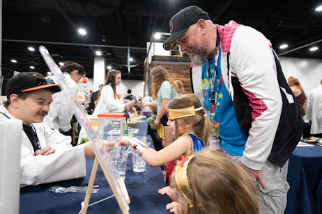 Two young girls, dressed as Wonder Woman, explore the Downtown Aquarium in the Pop Culture Classroom Kids' Lab. Their father looks on with joy, wearing a Ghost Spider sweater
