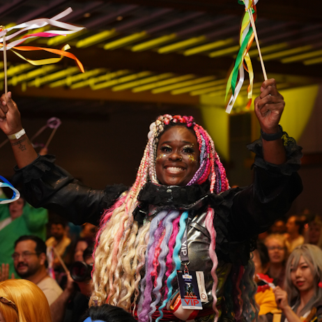 a fan smiles at the camera as she waves ribbons in celebration from the crowd. Her hair is braided, and it multiple colours like blue, purple, pink, and blonde.
