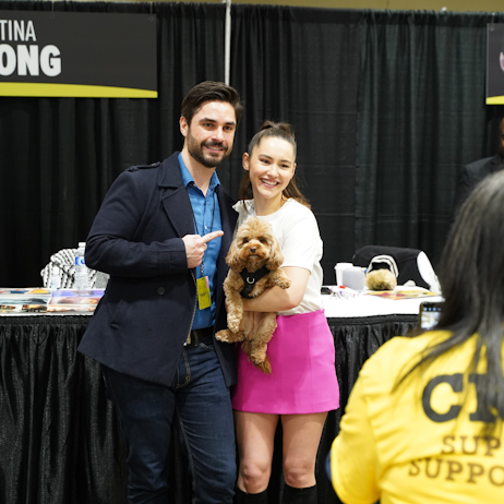 Christina Chong poses with a fan. He is in a navy blue plazer and light blue shirt with blue jeans. She is wearing a white tshirt and pink skirt with her hair in a ponytail. Between them, she holds her small brown puppy, Runa.