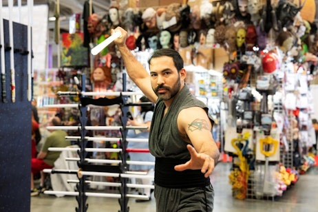 The image shows a man at a pop culture convention, actively posing with a lightsaber in a dynamic, action-oriented stance. He is wearing a sleeveless gray tunic, highlighting his muscular arms and a visible tattoo on his left arm. The backdrop is a vibrant and busy vendor's booth filled with a wide array of masks and pop culture memorabilia. The man’s focused expression and the motion blur on the lightsaber add a sense of energy and engagement to the scene, capturing the lively atmosphere of the event.