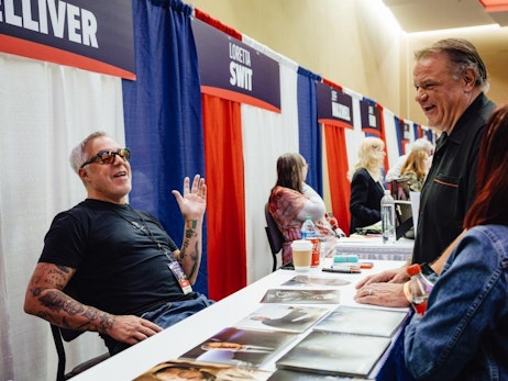 Two men engaging in a friendly conversation at a convention booth. The man on the left is Titus Welliver, with tattoos and sunglasses, sits behind a table with photographs, gesturing with his hand and smiling. The man on the right, wearing a black shirt, leans on the table and laughs. In the background, other attendees and booth staff are seen interacting.