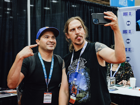 A man (left), smiling and pointing at Jason Mewes to his right sticking his tongue out and holding a phone for a selfie.