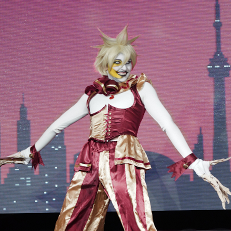 A fan, cosplaying as Mr. Sun from Five Nights At Freddy's: Sister Location strikes a pose on the main stage. Behind them is the Toronto Skyline in black on a red background. They are wearing a white long sleeve shirt with a half red and half gold corset, and gold and red striped pants.