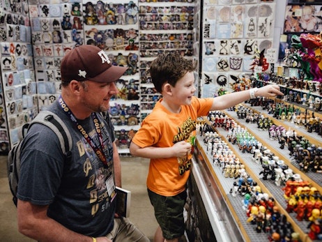 A man wearing a cap and a young boy in an orange shirt excitedly explore a convention booth. The boy, with wide eyes and a bright smile, points enthusiastically at a display of miniature figures, while the man looks on with interest. Behind them, a wall is covered with various stickers and drawings, adding to the vibrant atmosphere of the event.