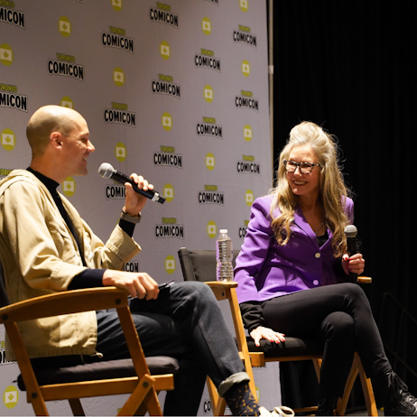 Mary McDonnell, in a purple blazer and black pants, leans in her seat to listen to the moderator, in a beige jacket and black pants, speak. Both are smiling.