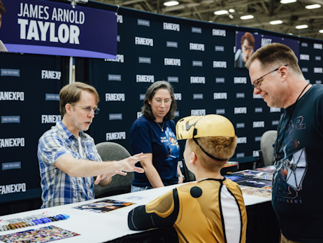 James Arnold Taylor (left) holding his hands out in a Jedi power pose, talking to a young boy and an older man (right)