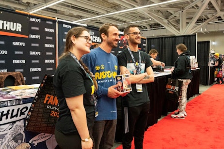 The image features three people standing together at a convention booth, smiling, and looking at something off-camera. They are located in a hall lined with various booths, indicated by background activity and a red-carpet aisle. The two men and one woman, all wearing casual attire and convention badges, seem engaged in a cheerful interaction. One of the men, voice actor Adam McArthur, is holding a Funko Pop! box, suggesting they might be discussing collectibles. The backdrop features the FAN EXPO banner prominently displayed, adding context to the event setting.