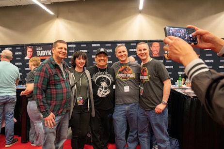 The image shows a group of four people posing for a photo with Danny Trejo at a convention booth with someone taking their picture. They are standing in front of a banner that features the FAN EXPO logo and images of celebrities. Three of the individuals are wearing matching 'Jurassic Park' T-shirts, suggesting they are fans or part of a themed group. The environment indicates a lively, casual setting typical of fan conventions, where attendees often meet celebrities or engage in fan-related activities. The atmosphere is cheerful and communal, capturing the essence of fan culture at such events.