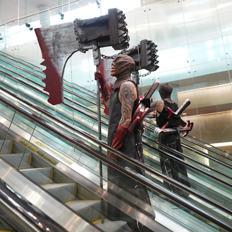 A creature cosplayer rides the escalators up, solo. Dawning a large weapon, half axe, half meat tenderizer. He is wearing a black smock and a bag over his head