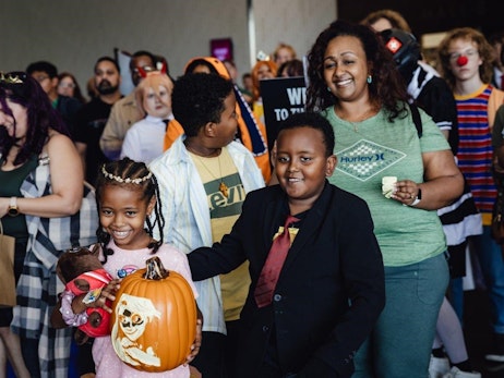 A joyful crowd gathers at an event. In the foreground, a smiling young girl with braided hair holds a large orange pumpkin decoration and a stuffed toy, while a young boy in a suit stands beside her, looking off to the side. A woman with a beaming smile stands behind them, wearing a green shirt and holding an item in her hand. The background is filled with other attendees, some in costumes, emphasizing the festive atmosphere.