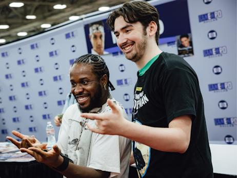 Shameik Moore (left) and a man (right), posing for a photo together holding their hands up in a Spider-Man web slinging pose.
