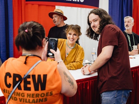 A woman in an orange "CREW" shirt, seen from behind, captures a moment on her phone where a long-haired man in a maroon shirt interacts with Alyssa Milano in a yellow blouse at a signing table. Behind them, two other men observe the scene. One wears a straw hat, and the other sports a dark shirt with a nametag. The setting appears to be an indoor event with a red and blue backdrop.
