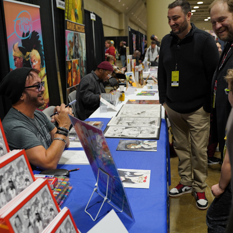 John Delaney, comic artist,  wearing dark glasses and a black beanie, sits at his table and chats with fans