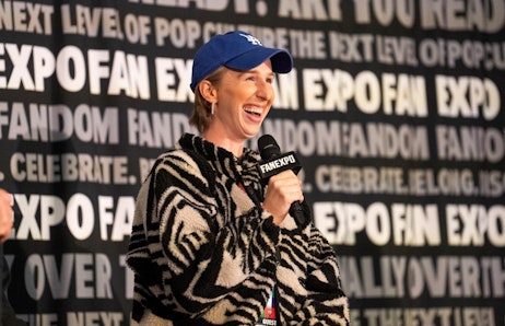 The image features Maggie Robertson with short hair, wearing a blue cap and a black-and-white zebra-striped jacket, speaking into a microphone at a FAN EXPO event. She is smiling broadly, conveying enthusiasm and joy as she engages with the audience. The background is a large banner filled with text related to the event, emphasizing its focus on pop culture and fandom. Her attire and casual style, along with her joyful demeanor, suggest a lively and interactive session, possibly involving fan interactions or a panel discussion.