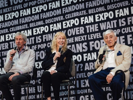 Three individuals sit on stage against a backdrop plastered with "FAN EXPO" and other related phrases. The man on the left, with silver hair and wearing a grey pullover, speaks into a microphone. In the center, a blonde-haired woman in a black blazer laughs joyously, also holding a microphone. To the right, an elderly gentleman in a beige jacket and blue pocket square listens attentively with a microphone in his hand. All are seated and engaged in a panel discussion at what appears to be a pop culture event.