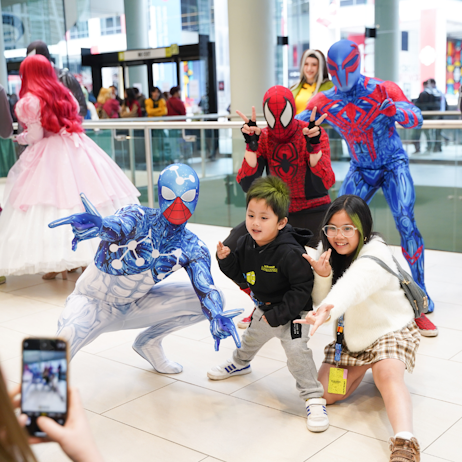 Two young children, middle and far right, strike the iconic spider-man pose with 3 spider-man cosplayers for their mother to take a photo