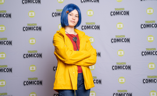 A girl dressed as Coraline stands confidently, her arms are crossed and she has the button key around her neck.