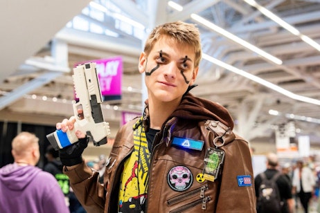 The image shows a young man at a convention, confidently posing with a futuristic-looking toy gun. He is dressed in a brown leather jacket adorned with various patches, including one with a yellow radiation symbol, and his shirt underneath displays vibrant comic book-style artwork. His face is marked with black paint resembling combat stripes, adding to his adventurous outfit. The background, filled with other attendees and neon lights, suggests a bustling, energetic environment typical of fan conventions. His expression and stance suggest he is engaged in a playful, action-oriented character portrayal.