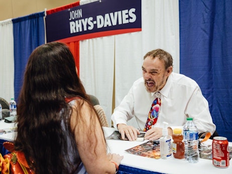 A man seated at a convention booth labeled "JOHN RHYS-DAVIES" enthusiastically engages with a visitor. The man, wearing a white shirt and a colorful tie, laughs heartily while talking to a woman with long brown hair, whose back is to the camera. On the table in front of him are various items including photographs, beverages, and a hand sanitizer bottle. The setting appears to be an indoor convention or fan event.