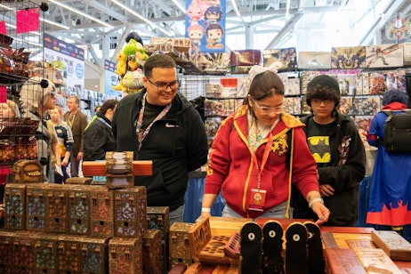 The image captures a vibrant moment at a comic con booth. Three young attendees are seen examining various intricately designed wooden boxes on a display table, which are likely merchandise or collectibles. The young man to the left is wearing glasses and smiling, observing a woman in the center, who is dressed in a red hoodie and appears focused on the items. Another youth, wearing a Batman shirt under a dark hoodie and a knitted beanie, stands to the right, also looking at the merchandise. The background is bustling with other attendees and a colorful array of additional merchandise, enhancing the lively atmosphere typical of such fan conventions.