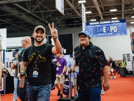 Two men walking into the show, smiling and holding up a peace sign, with a FAN EXPO Dallas banner in the background.