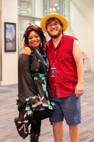 The image shows a joyful moment at a convention with two attendees posing for a photograph. The person on the left, a woman, is wearing a black layered dress with teal and white accents, and her hair is styled in vibrant red with streaks of black. She is also sporting a playful makeup look and a wide smile. The person on the right, a man, is dressed casually in a red sleeveless shirt and denim shorts, complemented by a straw hat. He is wearing glasses and also has a cheerful smile. The backdrop suggests they are inside a well-lit convention center, capturing the lively atmosphere of the event.