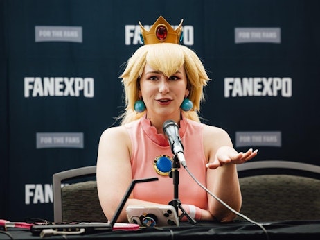 A woman in a cosplay outfit, resembling a character with a golden crown and large turquoise earrings, speaks into a microphone at a FANEXPO event. She wears a pink sleeveless dress with a blue emblem on it and gestures with her hand. The backdrop displays the "FANEXPO" logo repeatedly. On the table in front of her are various items, including a smartphone and some papers. She exudes confidence and engagement as she addresses her audience.