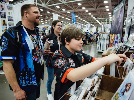 A family looking through art prints at a booth. There is a man on the left, a woman on the right, and a young boy in the front looking at the prints.