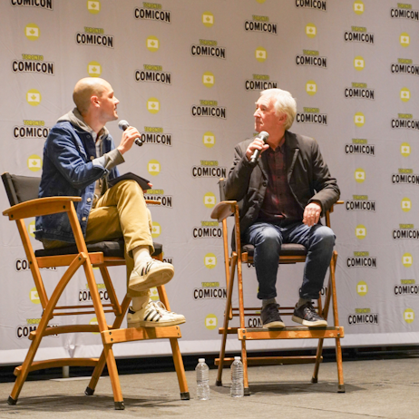 Denis Lawson (right) in a grey jacket, blue jeans, and red and green flannel shirt, sits and listens attentively to the moderator (wearing khaki pants and a denim shirt) as he asks a question.