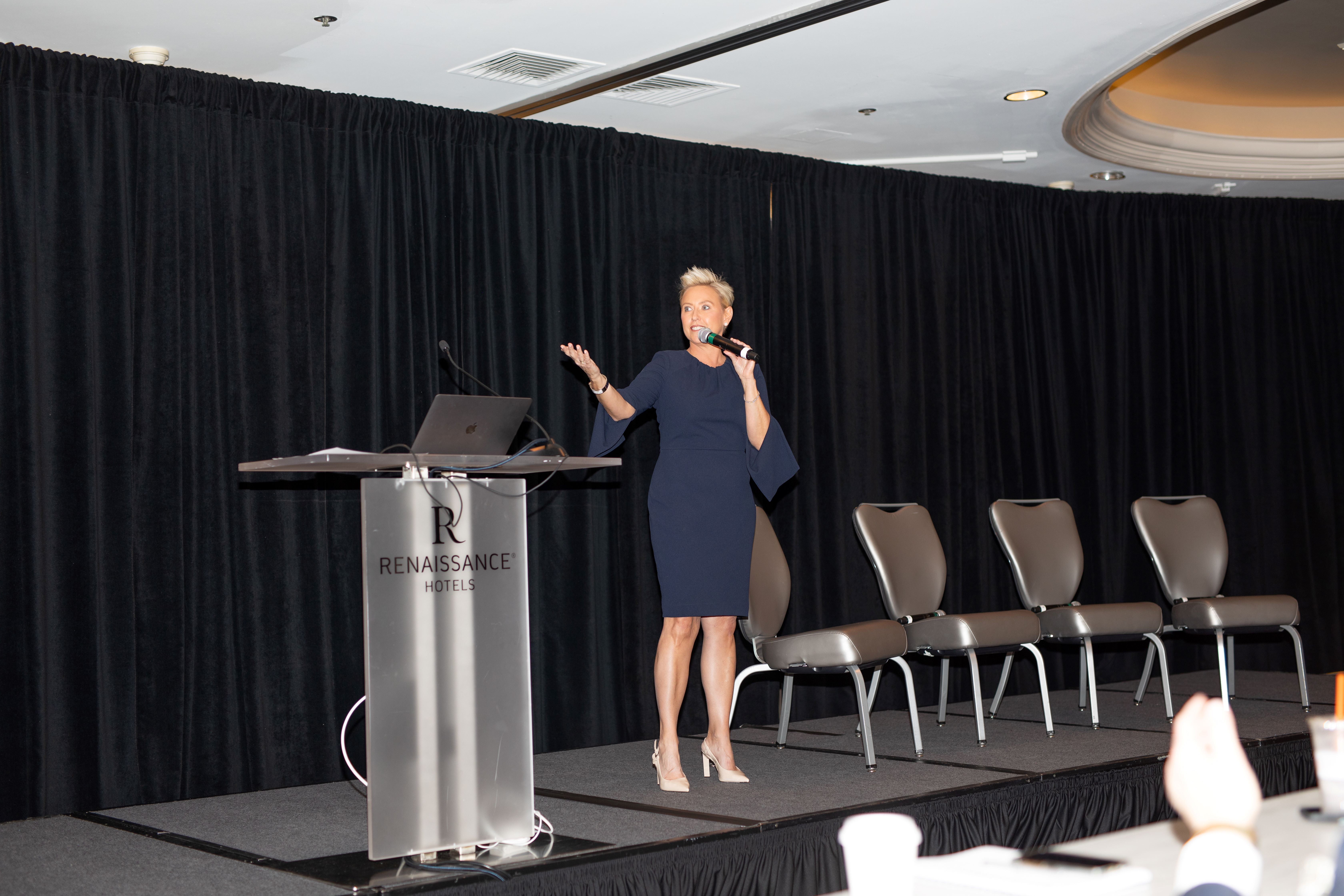 A spacious room with individuals seated at table listening to a keynote speaker.