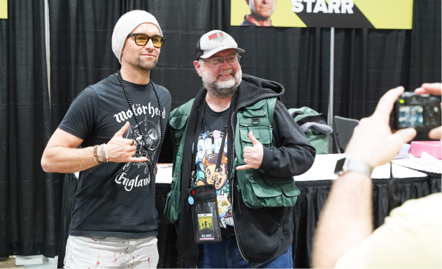 Antony Starr poses with a fan while a person only partially on camera (right) takes a photo with a digital camera