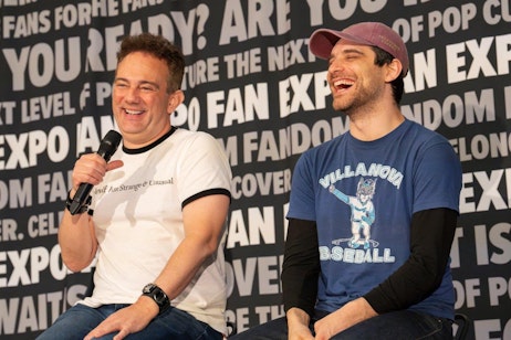 The image features Mike McFarland and Jeff Ward sharing a joyful moment on a panel at FAN EXPO. Mike, on the left, wearing a white t-shirt that reads "Myself Am Strange & Unusual," holds a microphone and laughs heartily, while Jeff, on the right, in a blue Villanova baseball t-shirt, also laughs, his eyes squinting in amusement. They are seated against a backdrop prominently displaying the FAN EXPO logo along with phrases like "Are you ready?" and "Ultimate fan experience," setting a lively and engaging atmosphere for the discussion. This scene captures the lighthearted and friendly interactions that are a hallmark of fan conventions.