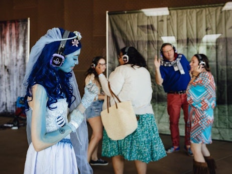 A woman with blue skin and long blue hair, wearing a white dress and veil, listens intently through purple headphones labeled "PARTY". She stands in the foreground, capturing attention with her distinct appearance. In the background, a group of people, including a man and a woman, also wear headphones and are engaged in a conversation. The setting appears to be an indoor event with draped fabrics and various attendees.