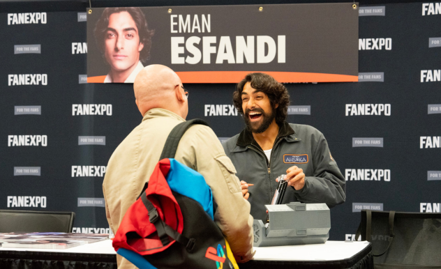 The image captures a joyful interaction at a FAN EXPO booth featuring Eman Esfandi, identified by the backdrop as "Eman Esfandi." He is engaging enthusiastically with a fan, and both share a laugh. The actor is dressed casually in a gray jacket with a patch labeled "Ahsoka," suggesting his role in a related franchise. He's signing an item for the fan, who is seen from the back, wearing a light-colored jacket and carrying a colorful backpack. The atmosphere is lively and fan-friendly, set against a backdrop with the actor’s promotional photo and event branding.