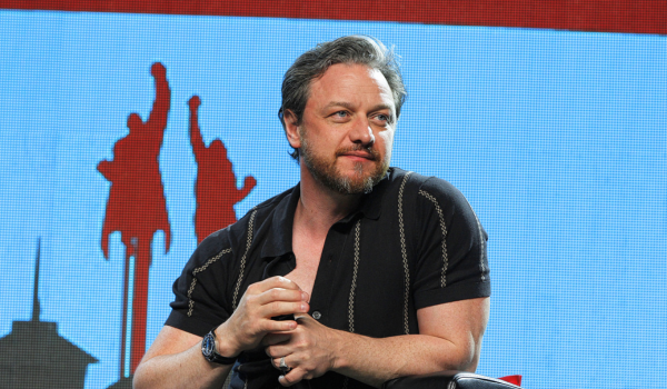James Mcavoy is sitting on stage in a black button up t-shirt with brown stripes. There is a blue and red background behind him. He is staring off into the crowd.