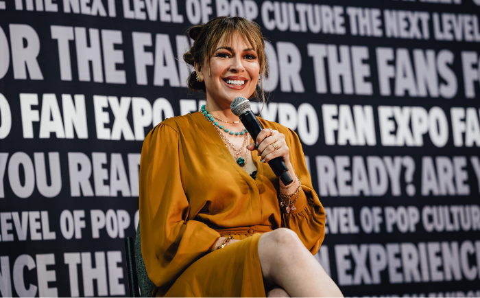 Alyssa Milano with shoulder-length brown hair sits in front of a backdrop displaying fan expo text. She dons an ochre dress with jewelry, including a turquoise necklace. Holding a microphone, she smiles warmly, showing her engagement in a conversation.