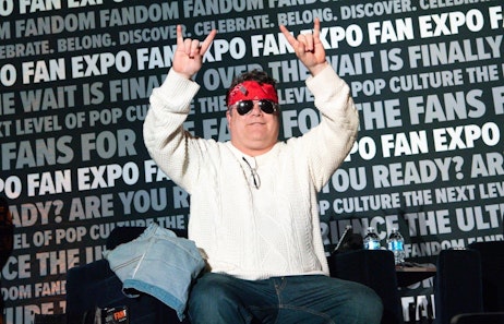 The image features Sean Astin on stage at a FAN EXPO event, joyously celebrating with his arms raised and making the "rock on" hand gesture. He is dressed in a white cable knit sweater and jeans, with a red bandana tied around his forehead. His expression is one of excitement and happiness. The background prominently displays the event's theme, with phrases like "FAN EXPO" and "ARE YOU READY?" in bold, suggesting a lively and engaging atmosphere typical of fan conventions.