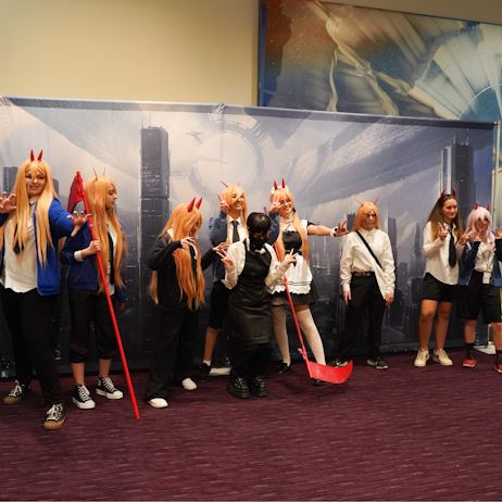 A Cosplay meetup for Chainsaw Man - each person is dressed as power. With black pants, a white button down, and black tie. All have the red devil horns, some have the matching scythe